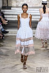 The image features a model walking down a runway wearing a stylish white dress. The dress has a fitted bodice with buttons down the front and a flowing, knee-length skirt. The skirt is accented with intricate lace detailing and a pink trim. The model has a sleek hairstyle and is wearing strappy sandals, adding to the elegant yet casual summer vibe of the outfit. The setting appears to be a fashion show with an audience in the background, emphasizing the runway atmosphere.