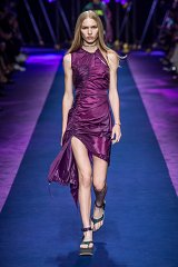 The image features a model walking down a runway, showcasing a striking purple dress. The dress has a unique design with ruching and a high slit, accentuating the model's silhouette. She is wearing stylish green platform sandals that complement the outfit. The runway is lit with vibrant purple and blue hues, adding an energetic atmosphere to the scene. The background shows an audience, although they are out of focus, highlighting the central focus on the model and her attire.