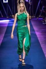 The image features a model walking on a runway wearing a vibrant green dress. The dress has a fitted, draped design with a side slit, emphasizing the model's silhouette. Her straight blonde hair is styled simply, complementing the overall look. The runway is illuminated with colorful lights, and the background suggests a high-fashion event. The setting creates a dynamic and stylish atmosphere.