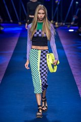 The image features a model walking down a runway, showcasing a vibrant fashion look. She is wearing a cropped top with long sleeves in various colors, including purple and green, and a checkered pattern. The bottom is a fitted skirt that combines bold green and black checkered designs. She carries a bright yellow handbag, and her footwear consists of chunky black sandals. The runway is illuminated with colorful lighting, creating a striking and energetic atmosphere.
