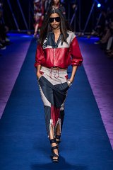 The image features a model walking down a runway during a fashion show. She is wearing a stylish outfit that includes a red and navy jacket made of leather, complemented by matching high-waisted pants. The ensemble has a sporty yet elegant vibe. The model also wears oversized sunglasses and strappy high-heeled shoes. The runway is lit with dramatic lighting, and the audience is visible in the background, creating an exciting atmosphere. The overall aesthetic is bold and fashionable, highlighting the designer's creative vision.