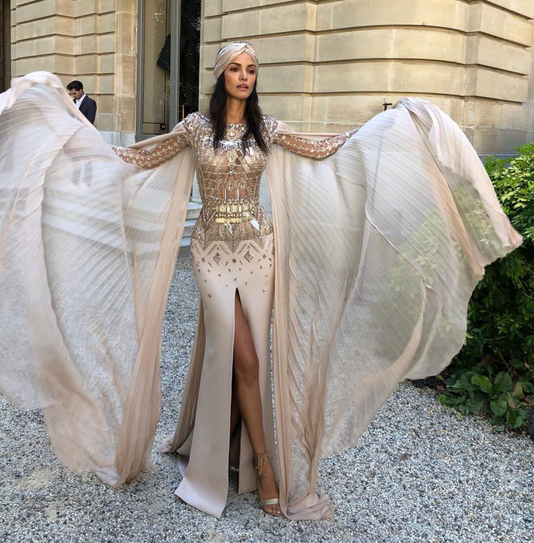 The image features a model wearing a stunning outfit that combines elegance and drama. The dress is a soft, neutral color with a fitted silhouette and a high slit on one side. The bodice is adorned with intricate beading and embellishments, giving it a glamorous look. Flowing, sheer cape-like sleeves extend from the shoulders, adding a sense of movement and grace. The model is also wearing a matching headpiece, which complements the overall style. The setting appears to be an outdoor area with architecture in the background, contributing to the sophisticated atmosphere.