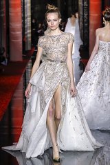 The image features a model walking down a runway in an elegant gown. The dress is intricately designed, showcasing a fitted bodice adorned with sparkling embellishments. It has a flowing, layered skirt that features a high slit, adding a dramatic flair. The gown transitions from a sheer upper section to a more structured bottom, enhancing its sophistication. The setting is a fashion show, with soft lighting and a reflective runway, contributing to the glamorous atmosphere. The model's hairstyle is sleek and minimalistic, complementing the opulence of the gown.